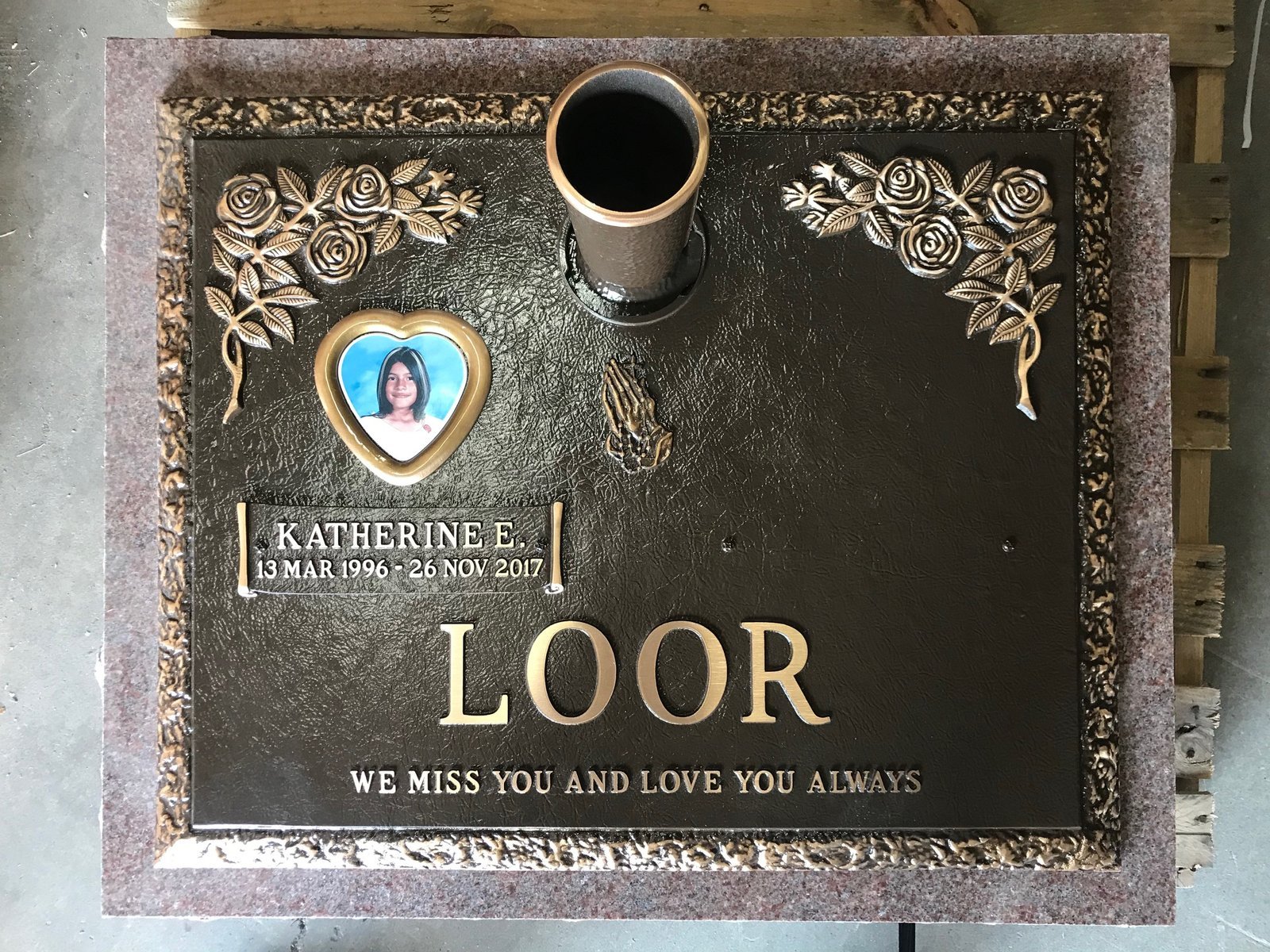 Bronze memorial marker on pink granite with rose corners, praying hands, and a heart portrait medallion