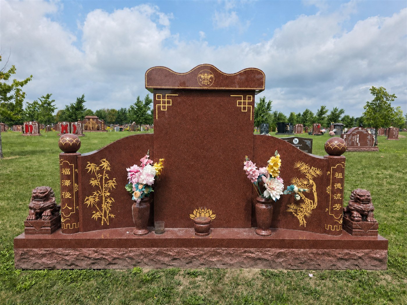 Red granite wing monument with a shaped cap, gold coloured bamboo and pine motifs and a lion figure at each end