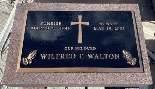 Bronze flat marker with a dark background, raised lettering, a plain cross and praying hands in the corners on pink granite