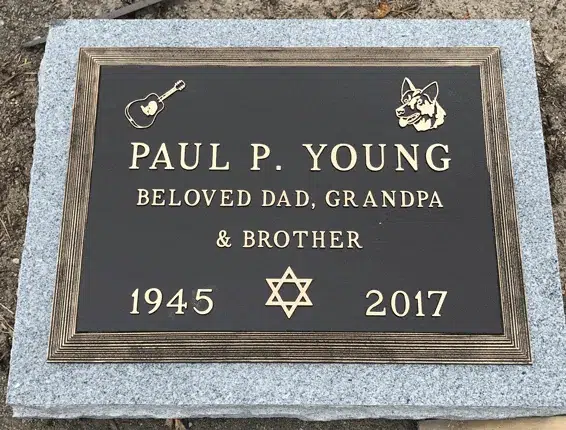 Bronze grave marker with an acoustic guitar, a dog head emblem and a Star of David between the years, on grey granite