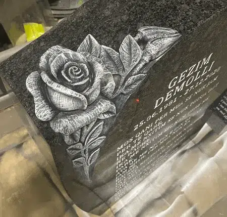 Rose carved in relief seen close up on the corner of a dark grey granite headstone with an engraved name, dates and epitaph