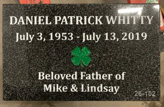 Dark granite flat grave marker with a painted green four leaf clover at the centre, a name, dates and a family dedication