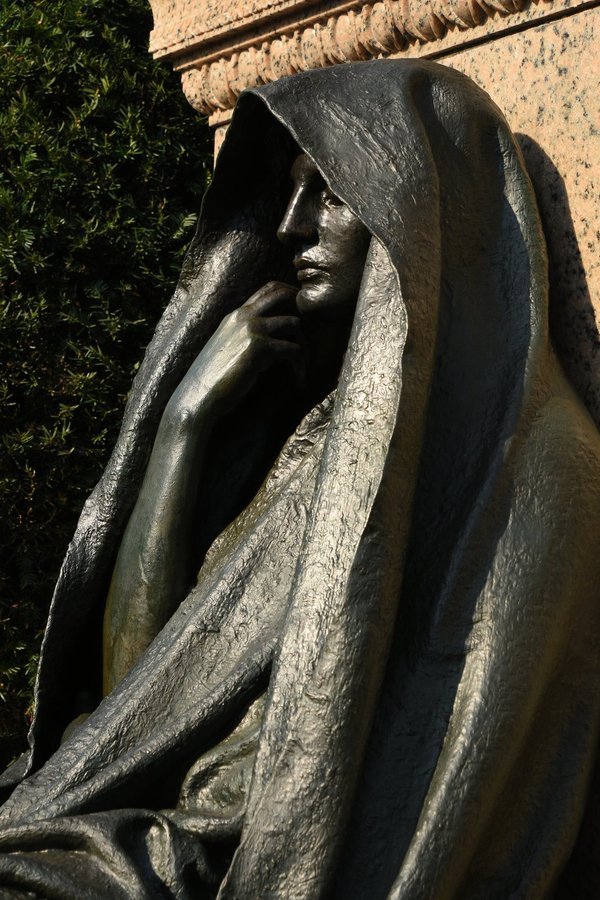 Close side view of a hooded bronze figure with a hand at its chin, pink granite behind, Rock Creek Cemetery, Washington DC
