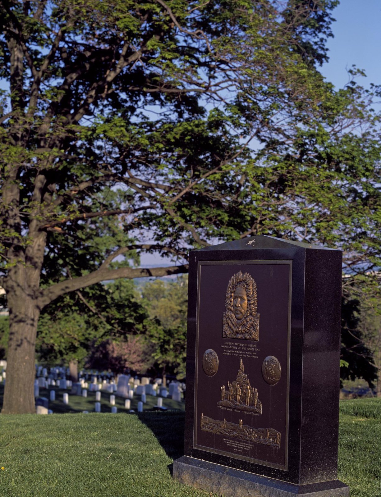 Polished black granite monument with a bronze relief plaque, rows of white headstones behind, Arlington National Cemetery