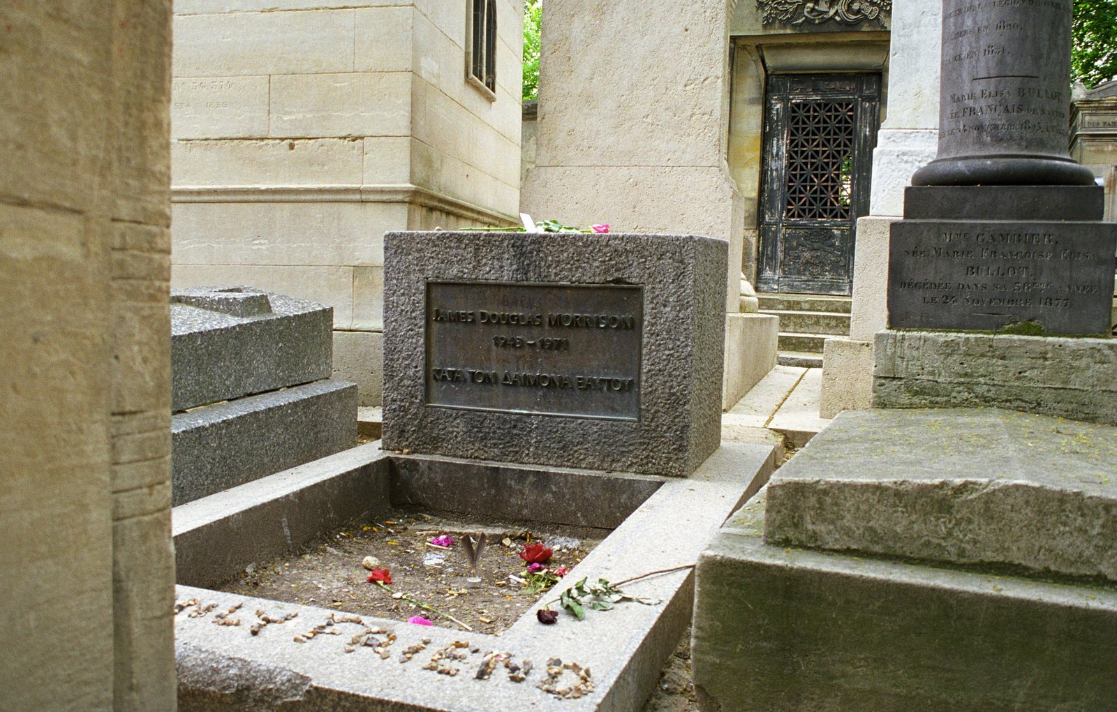 Grey granite block headstone with a bronze plaque above a bare plot strewn with roses at Père Lachaise Cemetery, Paris