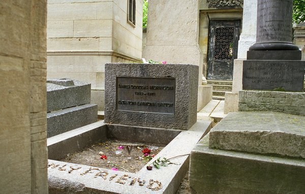 Rough grey granite block headstone with a bronze plaque, hemmed in by tall stone tombs at Père Lachaise Cemetery, Paris