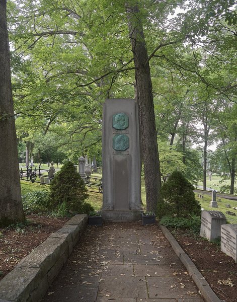 Tall grey stone shaft with two bronze portrait medallions under trees at Woodlawn Cemetery, Elmira NY