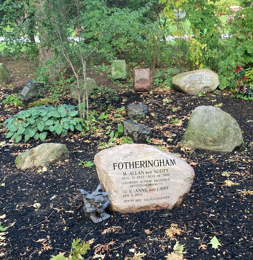 Engraved pink and grey boulder with a small gargoyle figure at its base, on dark mulch at Mount Pleasant Cemetery, Toronto