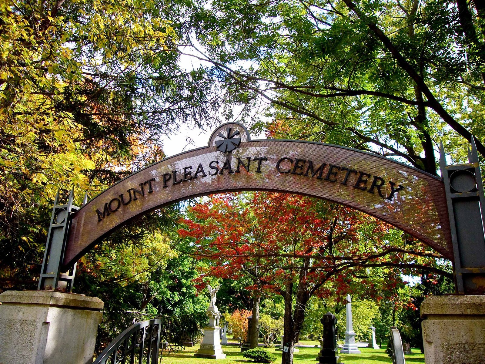 Arched metal sign on stone piers at an entrance to Mount Pleasant Cemetery, Toronto, with monuments and autumn trees beyond