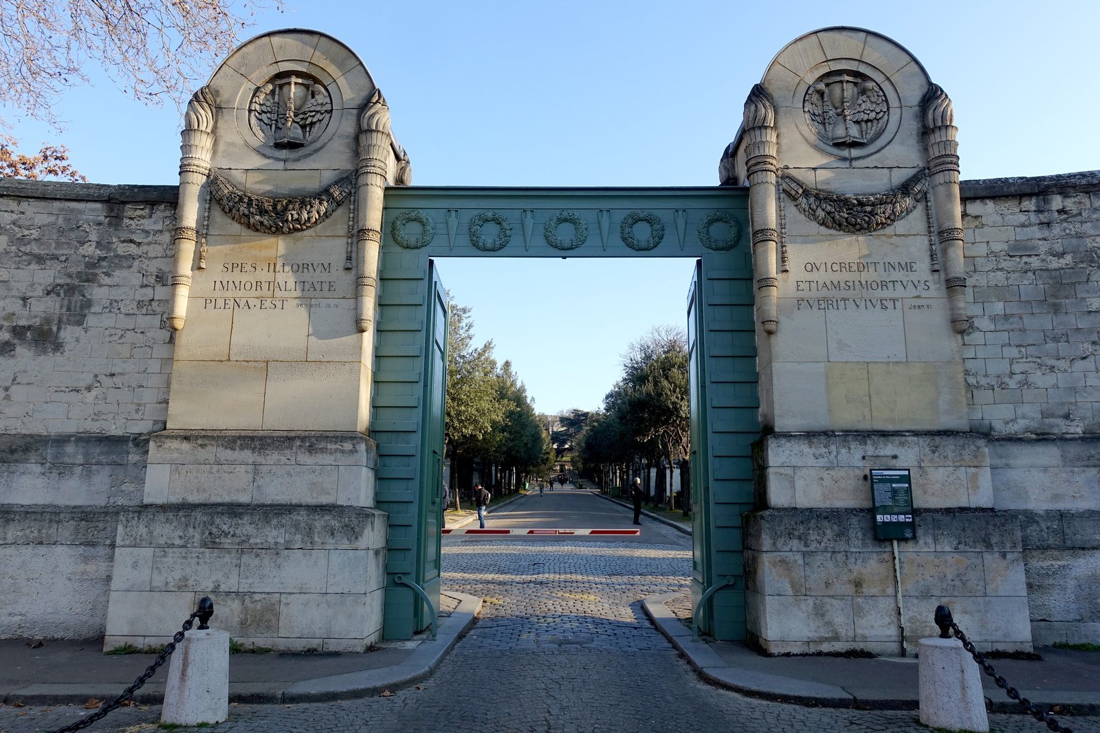 Entrance gate of Père Lachaise Cemetery in Paris, with carved pale stone pillars, open green gates and a tree lined lane