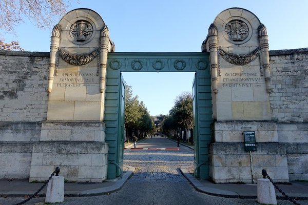 Carved stone entrance gate of Père Lachaise Cemetery in Paris, its green gates open to a tree lined lane