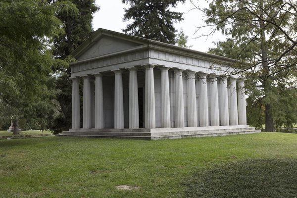 Pale stone mausoleum shaped like a Greek temple, with rows of fluted columns, on a cemetery lawn among trees