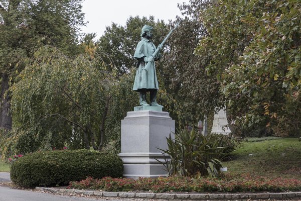 Green bronze soldier statue with a rifle on a tall grey granite pedestal, ringed by shrubs and red plants in a cemetery