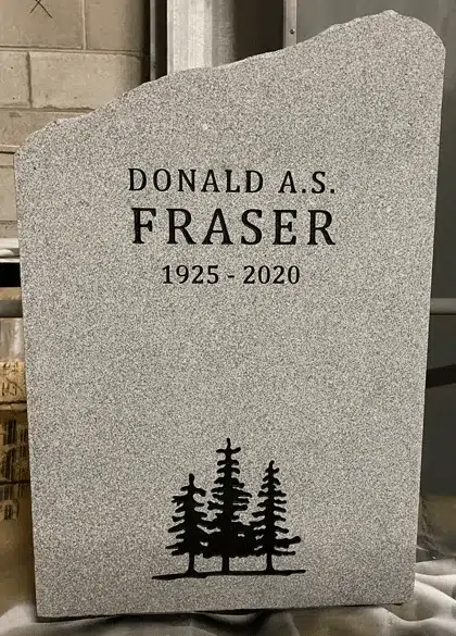 Light grey granite headstone with a rock pitched irregular outline, an engraved name and dates and three evergreen trees