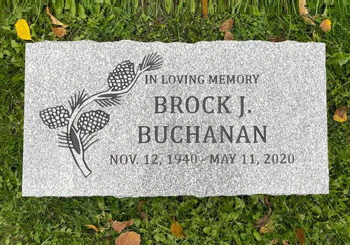 Light grey granite flat grave marker on grass with engraved pinecone and pine branch artwork beside a name and dates